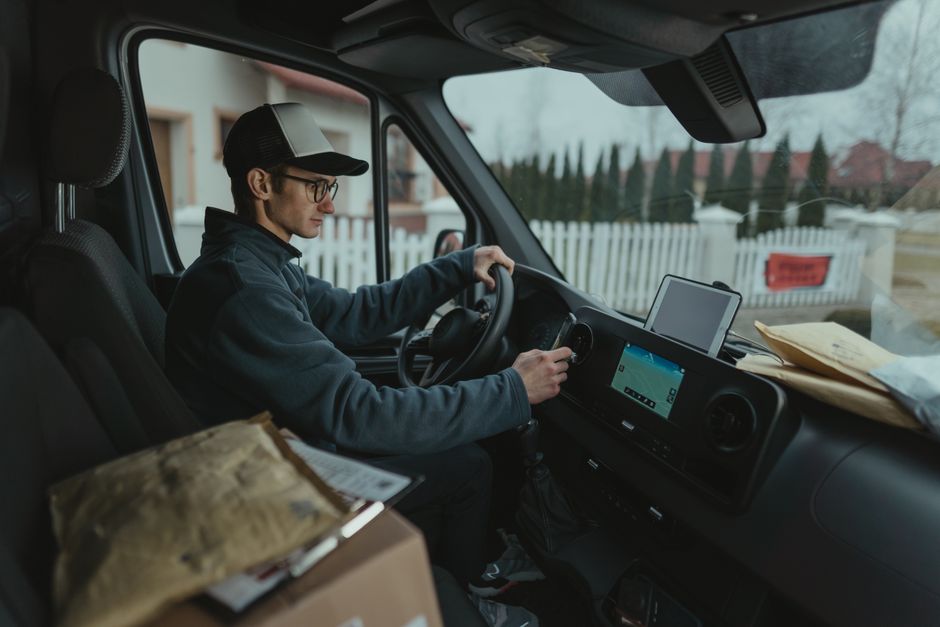 A silver delivery van parked on a suburban street during daylight hours, with a smiling driver sitting in the open window. The van displays the words 'DELIVERY ALWAYS ON TIME' on its side, indicating